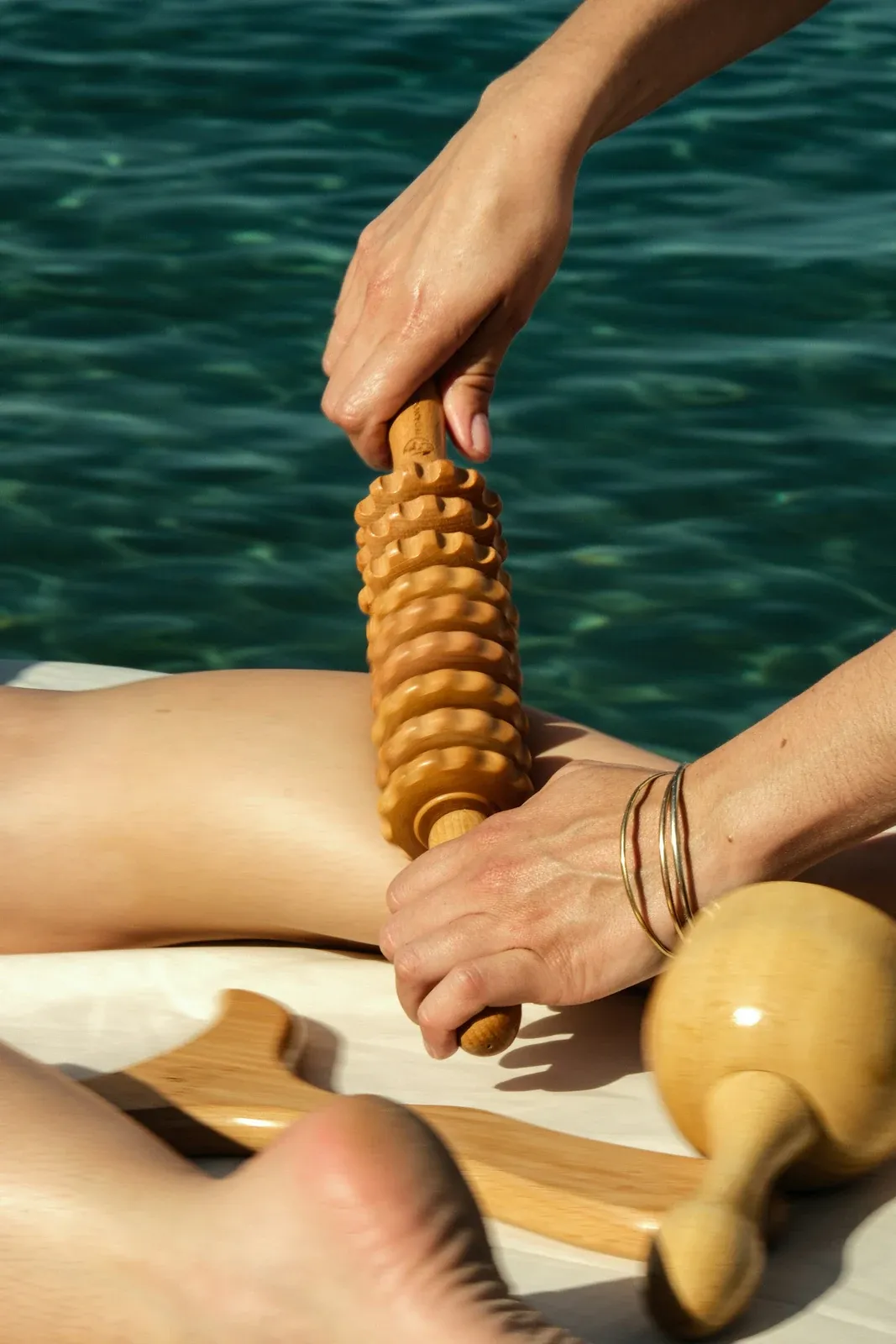 Mãos usando um rolo de massagem de madeira na perna, ao lado de uma piscina