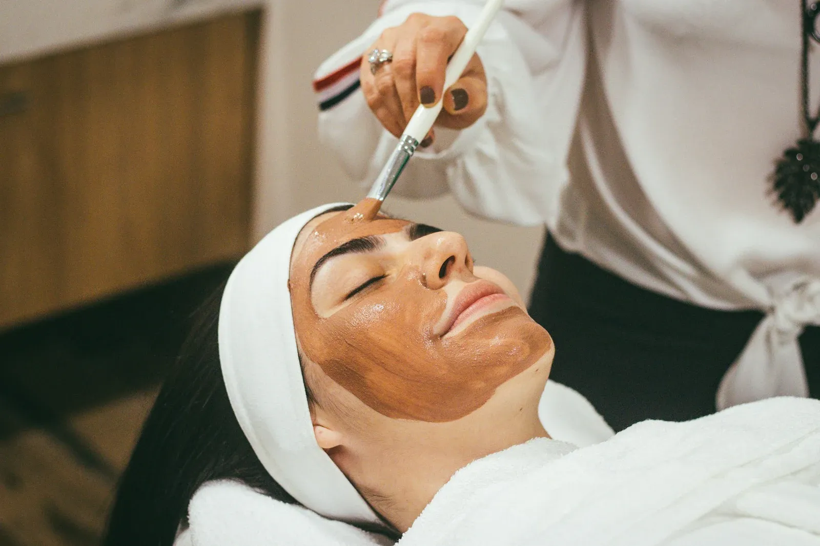 Mulher com faixa branca no cabelo recebendo máscara facial aplicada com pincel durante uma limpeza de pele