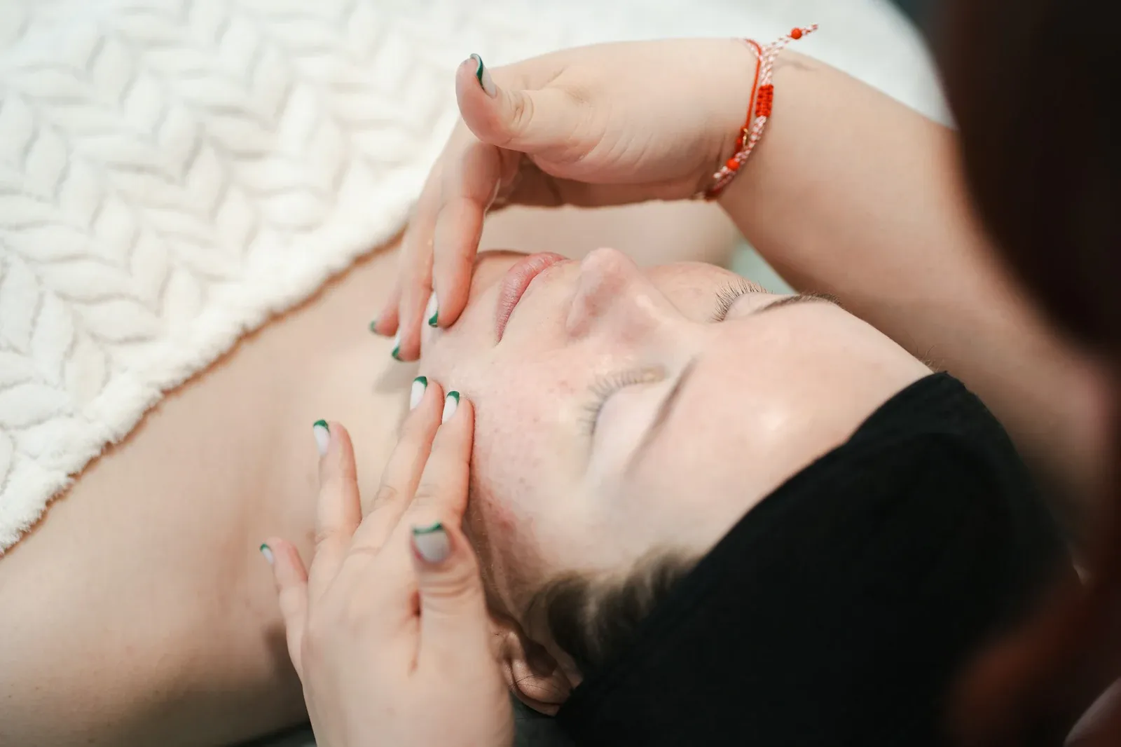 Mulher de olhos fechados recebendo um tratamento facial suave, com as pontas dos dedos da profissional nas têmporas